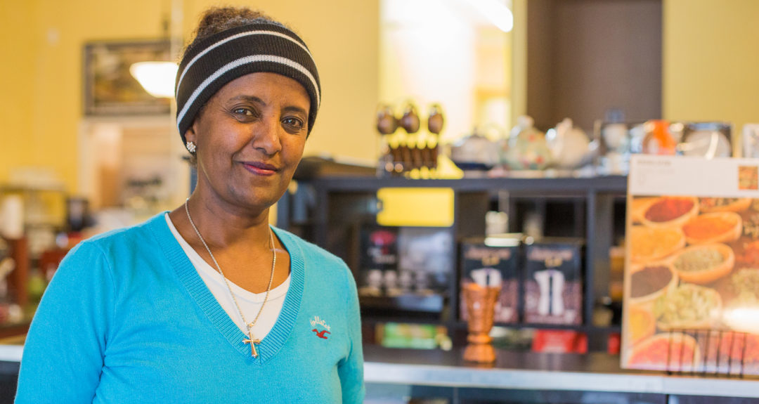 An Eritrean woman wearing a blue sweater stands in her market and restaurant.